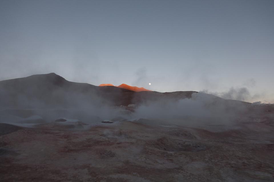 Steam moves slowly across the surface of the land.
The light arrives gradually, isolating the ridge in soft orange.
The rest of the field remains cold and grey.
The moon fades but does not disappear.