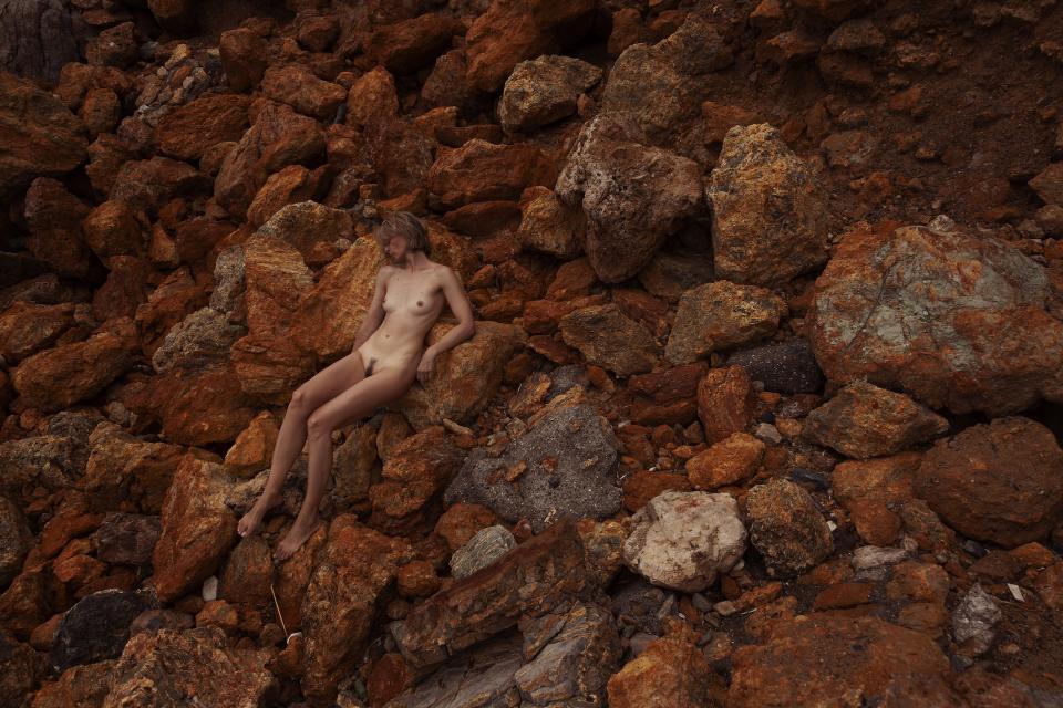 Large oxidized rocks fill the frame in layered formations.
A woman rests among them, her posture aligned with the incline.
The tones of her skin echo the surrounding mineral reds.
The distinction between body and landscape narrows.