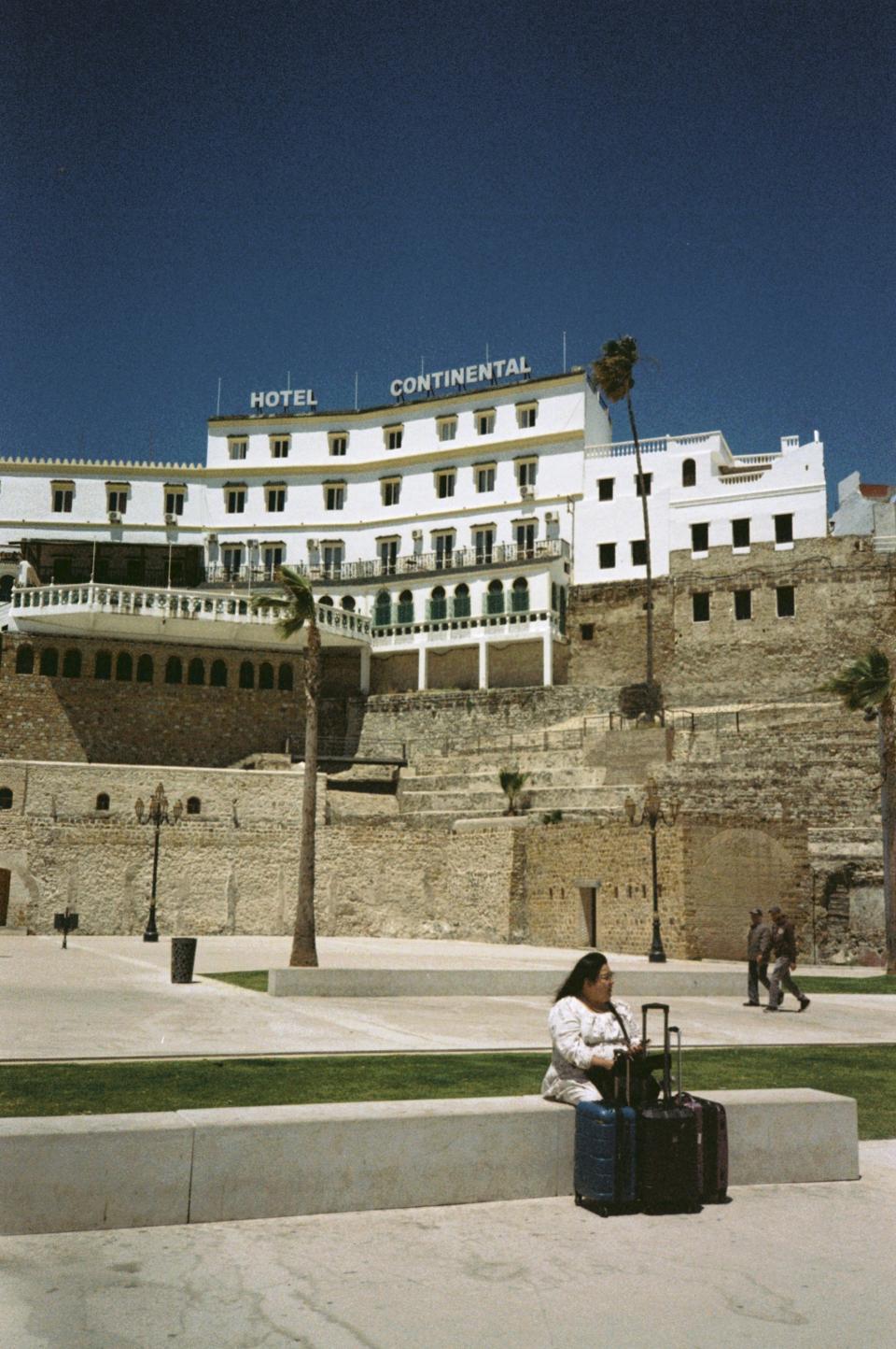 The white façade rises above layered stone walls, centered against a flat blue sky.
Palm trees lean slightly in the wind.
In the foreground, a woman sits on a low concrete bench with two suitcases beside her.
The building dominates the frame; she waits within it.
