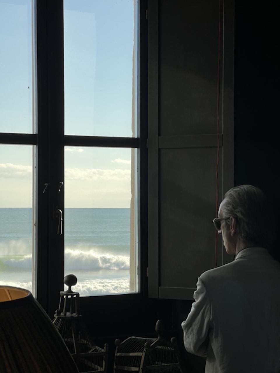 The window divides the view into four equal sections, the frame forming a cross.
Sea and sky occupy each quadrant without interruption.
The artist Felice Levini stands just off-center, facing outward.
Light outlines his profile against the moving water.