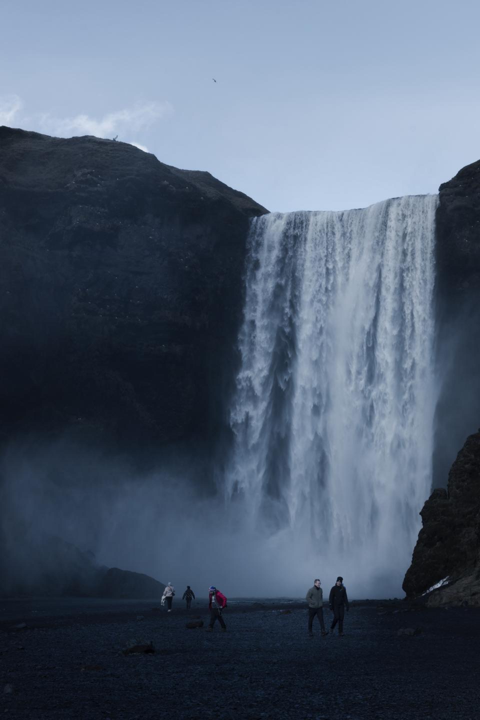 The cascade occupies most of the frame, uninterrupted from edge to edge.
Spray thickens the air near the ground.
A few people walk beneath it, spaced apart.
Their scale feels provisional.