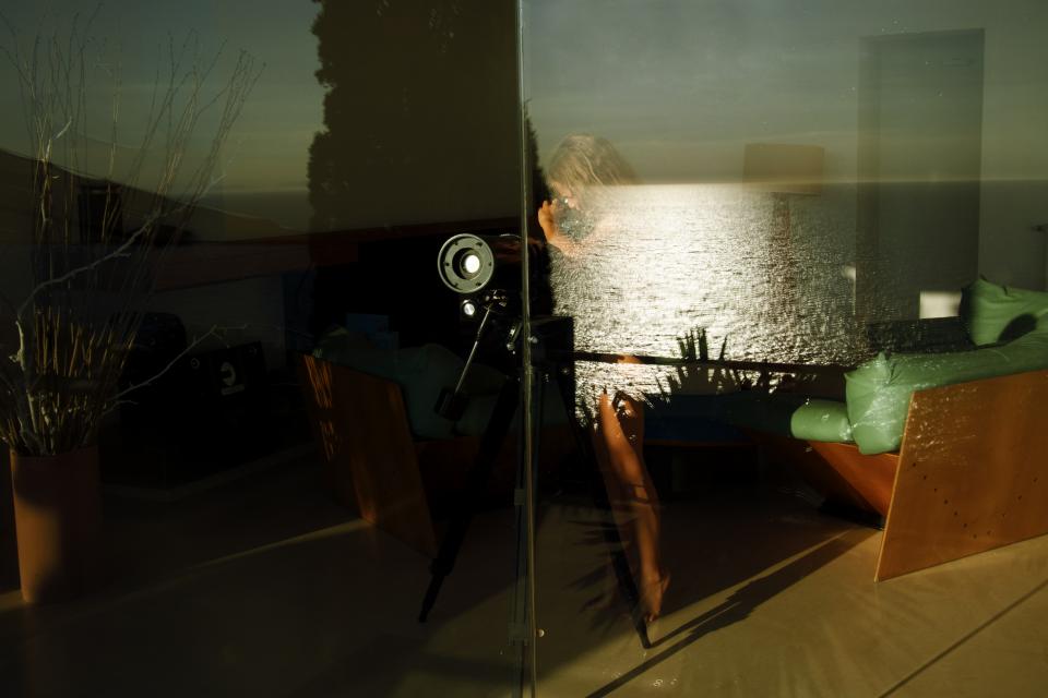 A vertical pane divides the room from the sea.
Outside, the horizon burns in late light.
A woman sits near the window, her figure partially absorbed by reflection.
The ocean overlays her body in shifting bands of gold.
Beside her, a telescope stands directed toward the water.
It suggests proximity without contact.
The glass keeps its distance intact.
The instrument narrows the view but does not cross it.
Interior and landscape occupy the same frame.
They do not touch.
The boundary remains transparent.
It holds.