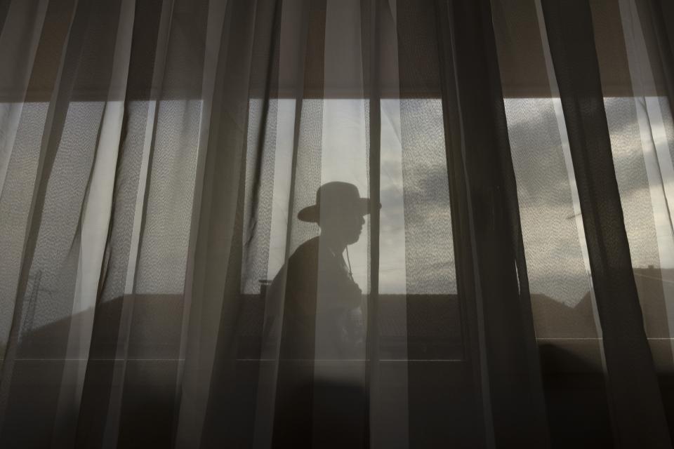 Through the curtain, a silhouette passes.
A hat cuts a sharp profile against the light.
The fabric becomes cover.
The camera stays low, partially concealed.
It feels like someone is hiding, waiting for danger to move on.
The figure outside continues past the window.
No contact.
Only distance.