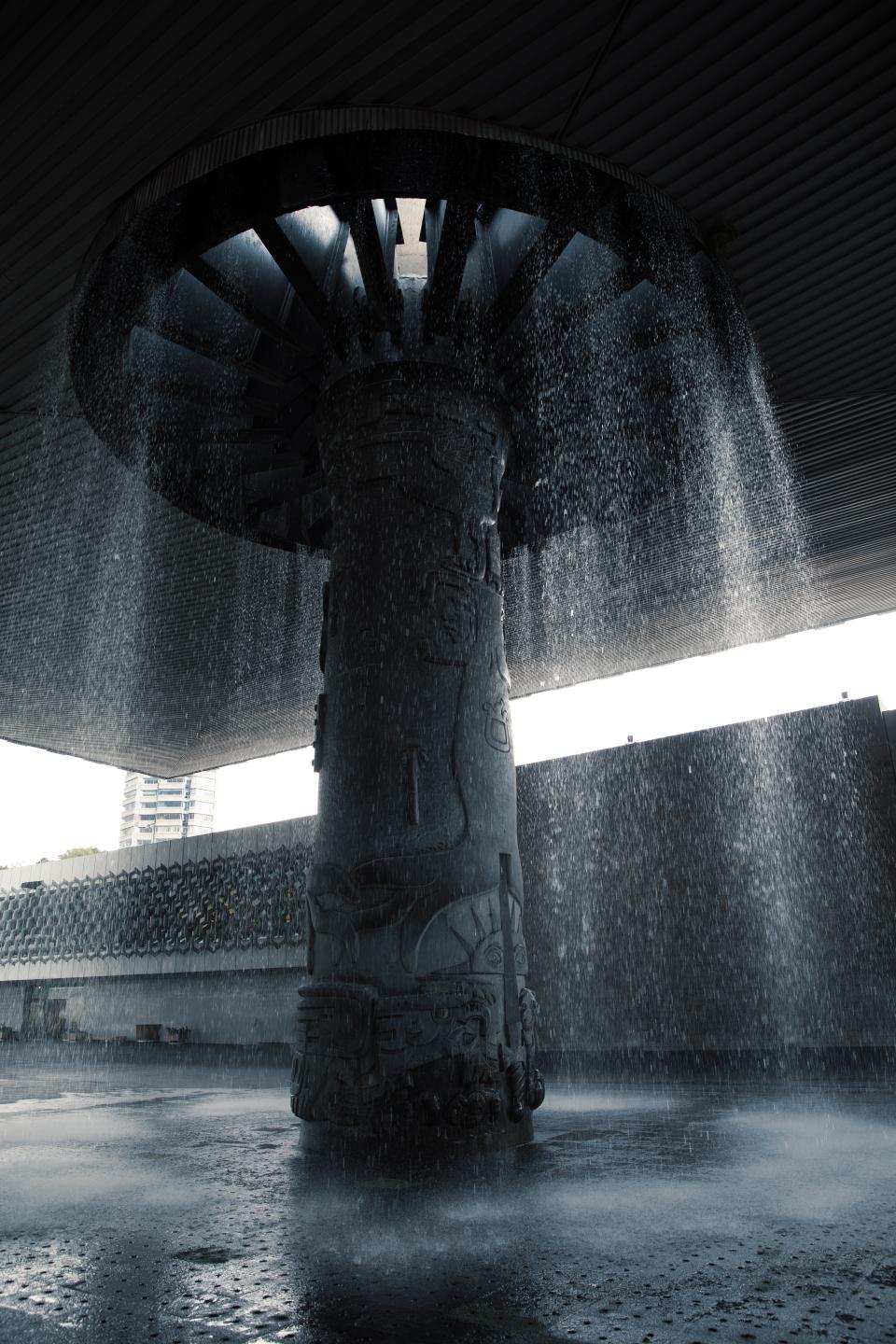 Water falls in a circular curtain from the concrete canopy above.
A single column rises at the center, carved with ancient forms.
Rain and architecture become one gesture.
The stone holds memory; the water erases and renews it at once.
Light filters through the opening overhead.
The past stands upright.
The present keeps falling.