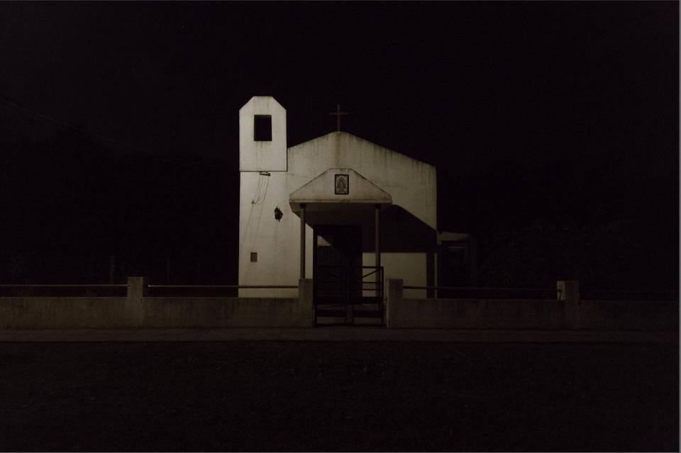 A white facade floats in darkness.
A red cross barely visible against the black sky.
The entrance is closed, unlit.
Shadows swallow the sides of the building.
No movement.