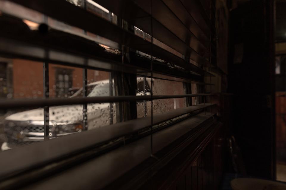 Blinds half-closed.
Water beads on the glass outside.
A white car sits across the street, blurred by rain.
The light is low, amber, interior.
The world is there — but filtered, segmented.
Nothing enters.
Nothing fully leaves.