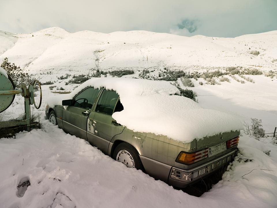 A car rests half-submerged in snow beside a wide, open field.
The accumulation on the roof is thick and intact.
The terrain beyond is uniform and pale.
No tracks lead away from it.