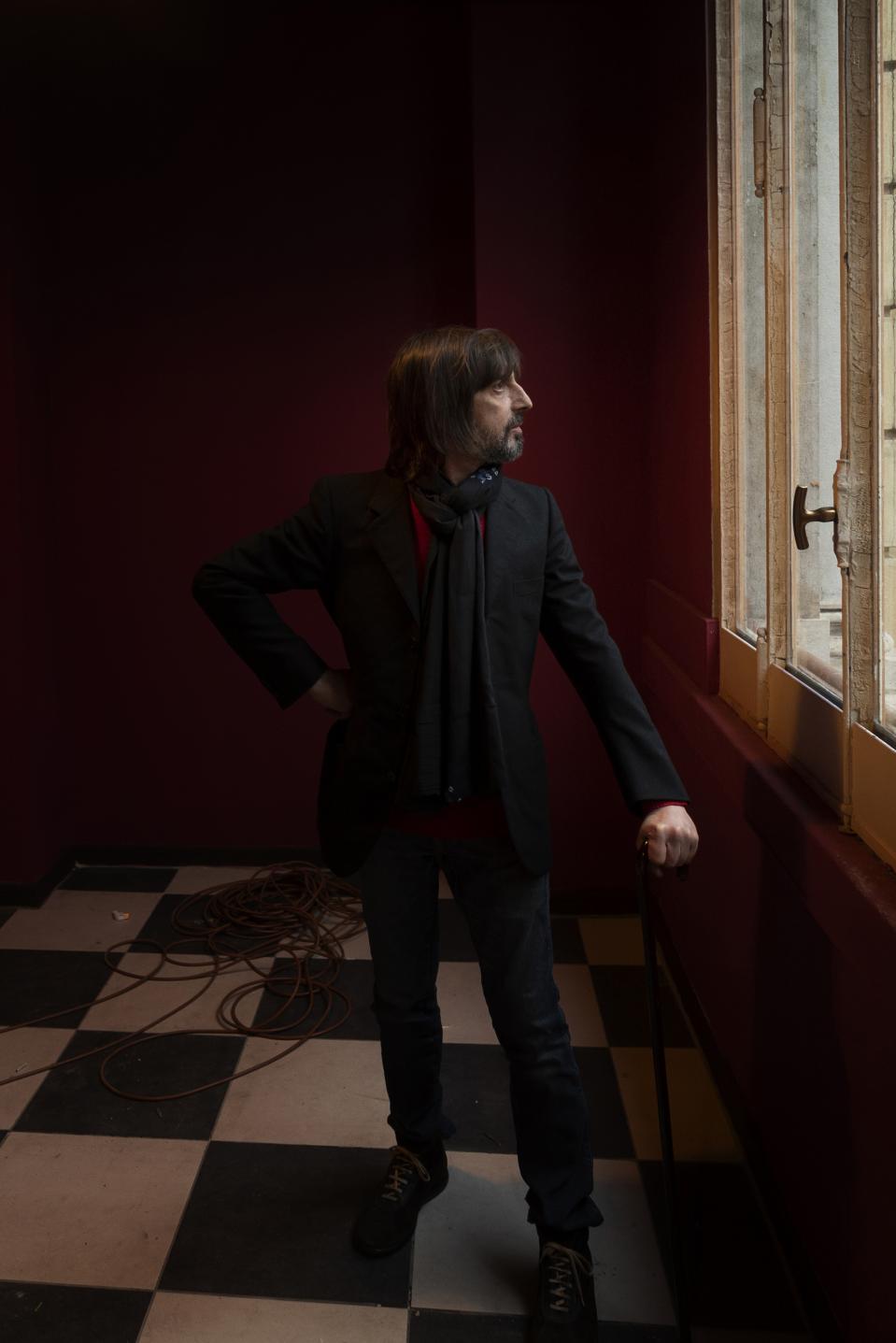 The artist Nicus Lucà stands beside the window.
One hand on his hip, the other resting on the sill.
The red walls absorb the light, intensify the silence.
Black jacket, dark scarf — a controlled presence against the saturated space.
The checkered floor fractures the ground beneath him.
Outside, something holds his attention.
Inside, nothing moves.